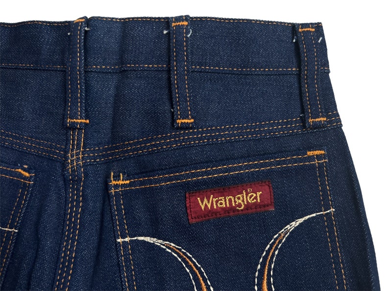 May include: A pair of dark blue denim jeans with orange stitching and a red leather Wrangler label on the back pocket.