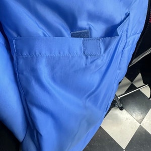 May include: Close-up of a light blue jacket interior with a rectangular pocket. The pocket is secured with a small velcro patch. The jacket's lining is a smooth, shiny material.