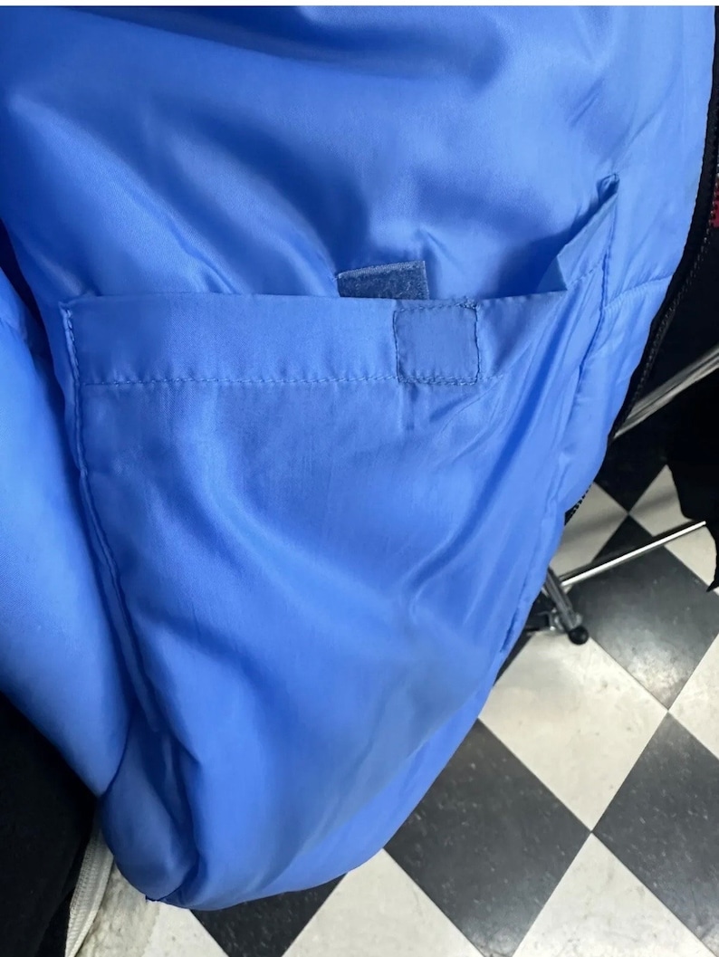 May include: Close-up of a light blue jacket interior with a rectangular pocket. The pocket is secured with a small velcro patch. The jacket's lining is a smooth, shiny material.