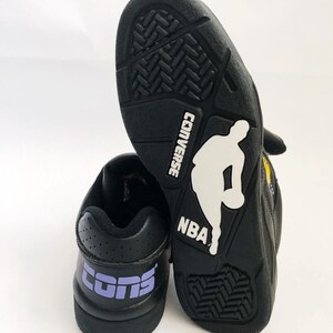 Puede incluir: Un par de zapatillas de baloncesto Converse negras con un logotipo "CONS" morado en la parte posterior de una zapatilla y una silueta blanca de un jugador de baloncesto con el texto "CONVERSE" y "NBA" en la parte posterior de la otra zapatilla.