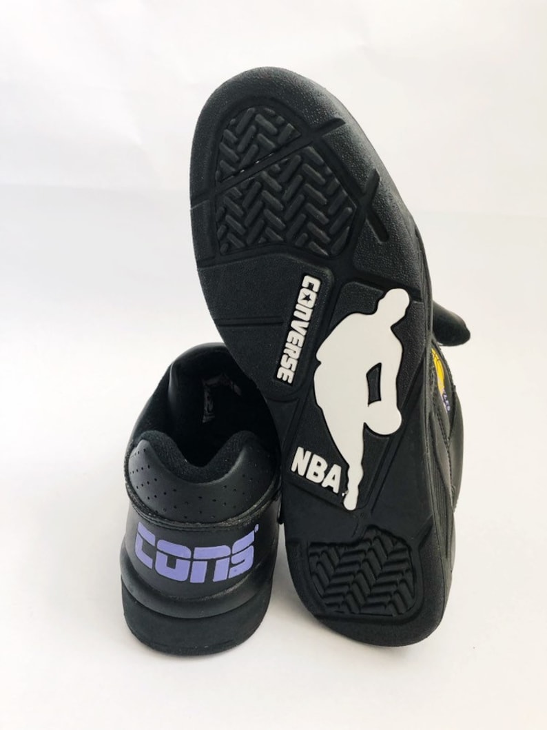 Puede incluir: Un par de zapatillas de baloncesto Converse negras con un logotipo "CONS" morado en la parte posterior de una zapatilla y una silueta blanca de un jugador de baloncesto con el texto "CONVERSE" y "NBA" en la parte posterior de la otra zapatilla.