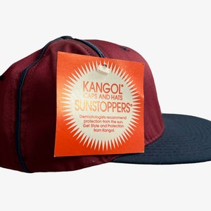 May include: A burgundy and black baseball cap with a Kangol tag attached. The tag is orange with a sunburst design and the text "Kangol Caps and Hats Sunstoppers". Dermatologists recommend protection from the sun. Get Style and Protection from Kangol.