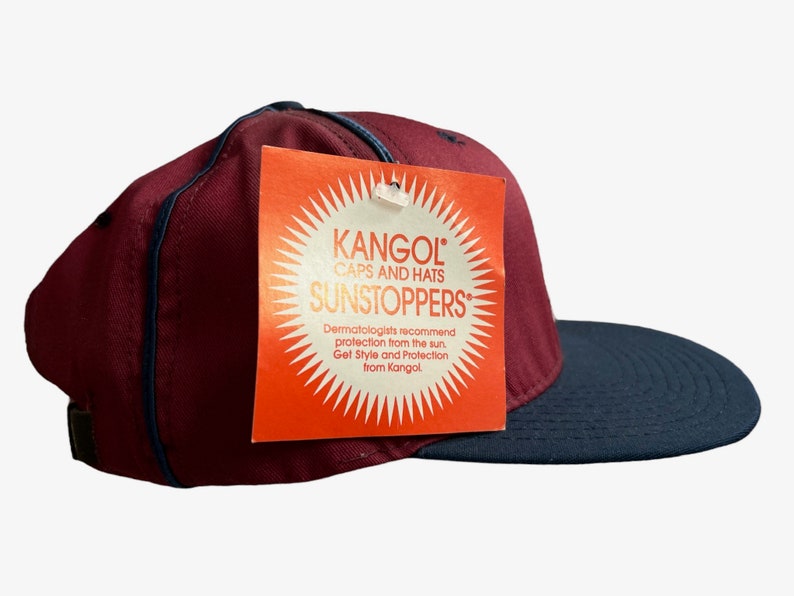 May include: A burgundy and black baseball cap with a Kangol tag attached. The tag is orange with a sunburst design and the text "Kangol Caps and Hats Sunstoppers". Dermatologists recommend protection from the sun. Get Style and Protection from Kangol.