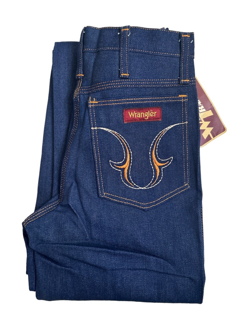 May include: A pair of dark blue denim jeans with a Wrangler label on the back pocket. The pocket has a decorative orange and white embroidery design.