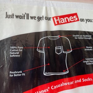 以下が含まれることがあります： Hanesのロゴとテキストが入った白いプラスチックパッケージ。パッケージには、ポケット付きの白いTシャツの図があり、二重襟、ゆったりとしたアームホール、防縮加工された綿などの特徴が強調されています。「Just wait'll we get our Hanes on you.」と書かれています。