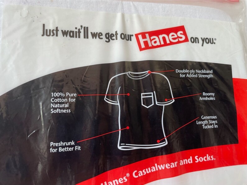 以下が含まれることがあります： Hanesのロゴとテキストが入った白いプラスチックパッケージ。パッケージには、ポケット付きの白いTシャツの図があり、二重襟、ゆったりとしたアームホール、防縮加工された綿などの特徴が強調されています。「Just wait'll we get our Hanes on you.」と書かれています。