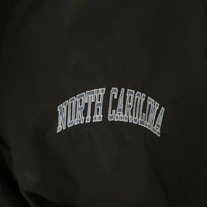 May include: Black jacket with the embroidered text "NORTH CAROLINA" in light blue. The lettering is arched across the chest. The jacket has a blue accent on the collar.