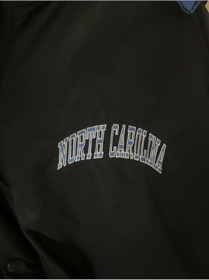 May include: Black jacket with the embroidered text "NORTH CAROLINA" in light blue. The lettering is arched across the chest. The jacket has a blue accent on the collar.