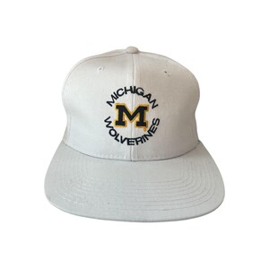 May include: A white baseball cap featuring the text "MICHIGAN WOLVERINES" in black and gold. The cap has a flat brim and a rounded crown. The logo includes a large gold "M" inside a black outline.
