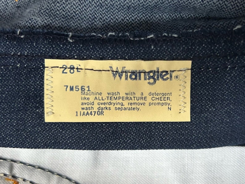 May include: A yellow care label with the text "Wrangler" and instructions for washing the jeans. The label also includes the size "28" and the number "7M561".