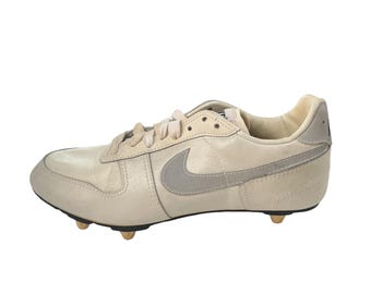 May include: A pair of off-white leather soccer cleats with a gray Nike swoosh logo. The cleats have a lace-up closure and a rubber sole.