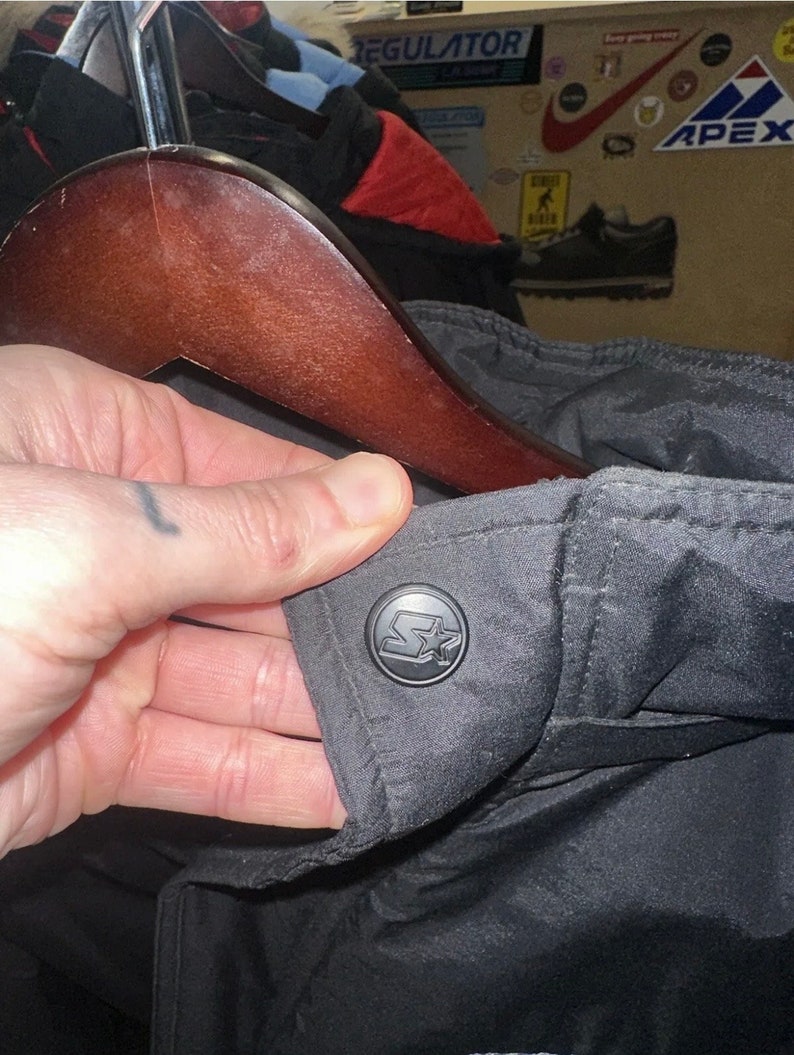 May include: Black jacket with a Starter logo button. The jacket is hanging on a wooden hanger. The Starter logo is a black circle with a white star and the letter "S".