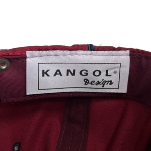 May include: A burgundy baseball cap with a white label that reads "Kangol Design" in black lettering.