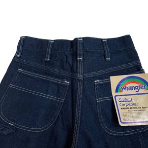 以下が含まれることがあります： 白いステッチと、後ろのポケットに付けられた Wrangler Student Carpenter Wrangler Utility Jeans のタグが付いた、濃い青色のデニムのカーペンタージーンズです。