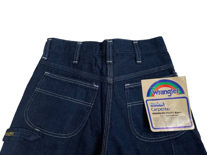 以下が含まれることがあります： 白いステッチと、後ろのポケットに付けられた Wrangler Student Carpenter Wrangler Utility Jeans のタグが付いた、濃い青色のデニムのカーペンタージーンズです。