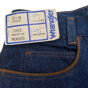 May include: A close-up of a blue denim jean with a brown leather trim on the pocket. The jean has a Wrangler tag attached with the text "Wrangler" and "Made in USA". The tag also includes the size "27 x 36 Student", style "134SPWR", and content "1295 100% Cotton".