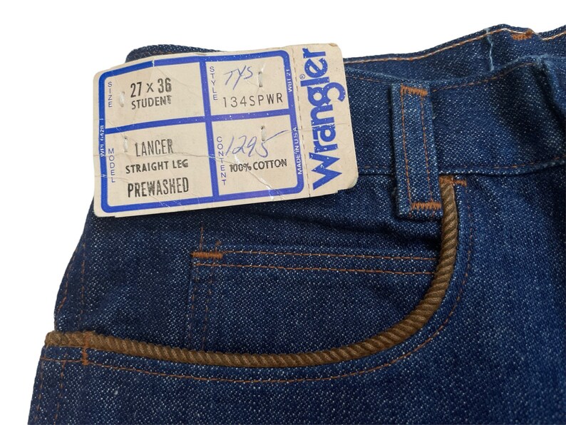 May include: A close-up of a blue denim jean with a brown leather trim on the pocket. The jean has a Wrangler tag attached with the text "Wrangler" and "Made in USA". The tag also includes the size "27 x 36 Student", style "134SPWR", and content "1295 100% Cotton".