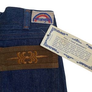 May include: Blue denim jeans with a brown corduroy patch on the back pocket. The patch has an embroidered design. A white tag with the text "FULL 1 YEAR WARRANTY" is attached to the jeans.