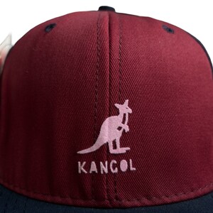 May include: A burgundy baseball cap with a black brim. The front of the cap features a pink kangaroo logo with the word "KANGOL" underneath.