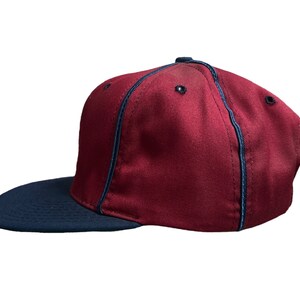 May include: A maroon baseball cap with a navy blue brim and a navy blue stitching detail around the crown.