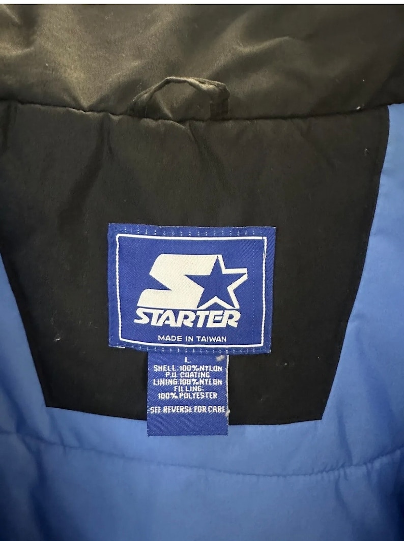 May include: A close-up of a Starter jacket label. The label is blue and white with the Starter logo and text. The jacket is black and blue. The label reads "MADE IN TAIWAN" and lists the materials: 100% Nylon shell, P.U. coating, 100% Nylon lining, and 100% Polyester filling.