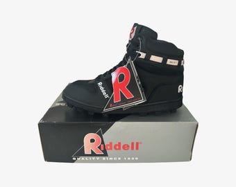 May include: Black Riddell football cleat with a high-top design, sitting on its original box. The cleat features the Riddell logo in white and red, with a tag attached. The box has the Riddell logo and the text "QUALITY SINCE 1929."