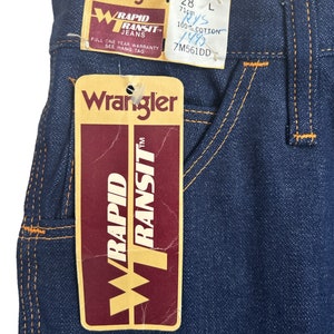 May include: A pair of blue denim jeans with orange stitching. The jeans have a Wrangler Rapid Transit label attached. The label has the text "Wrangler Rapid Transit Jeans" and "Full One Year Warranty See Hang Tag". The label also has the size 28, 71cm, L, 100% Cotton, 148, and 7M561DD.