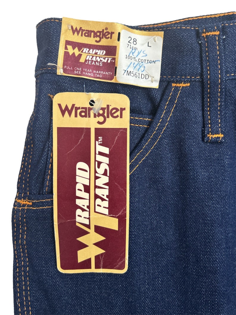 May include: A pair of blue denim jeans with orange stitching. The jeans have a Wrangler Rapid Transit label attached. The label has the text "Wrangler Rapid Transit Jeans" and "Full One Year Warranty See Hang Tag". The label also has the size 28, 71cm, L, 100% Cotton, 148, and 7M561DD.