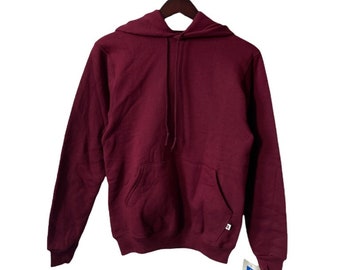 May include: A maroon hoodie with a drawstring hood and a kangaroo pocket. The sweatshirt is hanging on a wooden hanger. A white tag with blue and red text is attached to the sleeve.