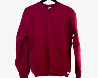 May include: A maroon crew neck sweatshirt hanging on a black hanger. The sweatshirt has a small white tag on the bottom right side.