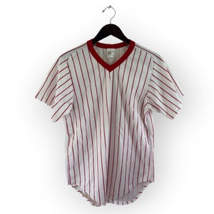 May include: White baseball jersey with red vertical stripes and a red V-neck collar. The short-sleeved shirt is hanging on a black wooden hanger. The jersey has a curved hemline.