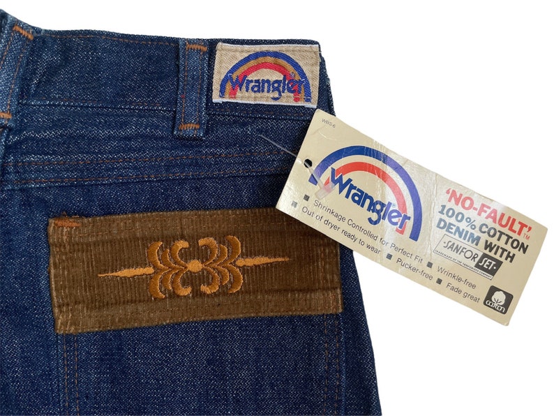 May include: A pair of blue denim jeans with a brown corduroy patch on the back pocket. The patch has an embroidered design. The jeans have a Wrangler label on the back waistband. The label says "Wrangler" and has a rainbow design. A tag attached to the jeans says "NO-FAULT" 100% COTTON DENIM WITH -SANFOR SET. Shrinkage Controlled for Perfect Fit Wrinkle-free Out of dryer ready to wear Pucker-free Fade great.