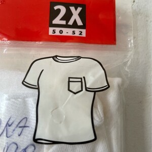 以下が含まれることがあります： ポケット付きの白いTシャツ、2XLサイズが透明なビニール袋に入っています。赤いタグにはサイズが黒で表示されています。Tシャツは袋の中で折りたたまれています。