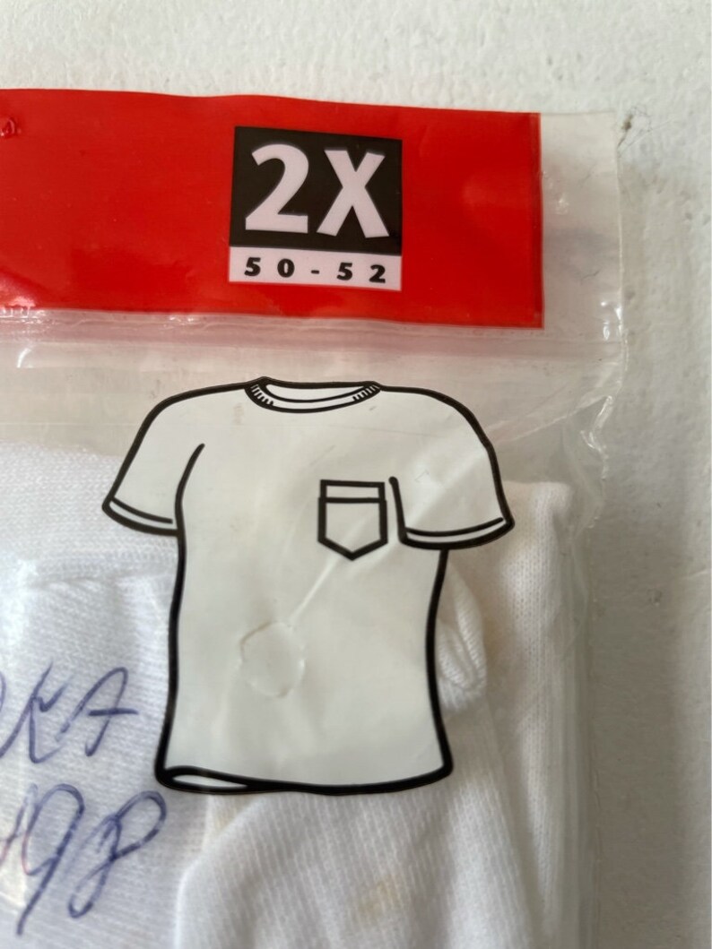 以下が含まれることがあります： ポケット付きの白いTシャツ、2XLサイズが透明なビニール袋に入っています。赤いタグにはサイズが黒で表示されています。Tシャツは袋の中で折りたたまれています。