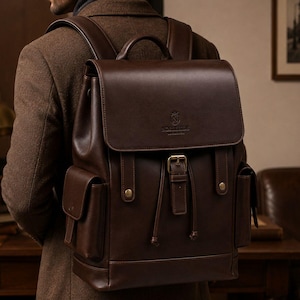 Può includere: Uno zaino in pelle marrone scuro con patta superiore dotata di chiusura con fibbia centrale, due tasche laterali e una maniglia superiore. La patta anteriore presenta un piccolo logo in rilievo con il testo "NOBLEHIDE" e uno stemma.