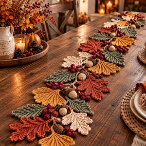 May include: A decorative table runner featuring crocheted autumn leaves in shades of rust, mustard yellow, cream, and sage green, interspersed with small crocheted acorns and clusters of dark red berries, arranged in a long, flowing seasonal pattern.