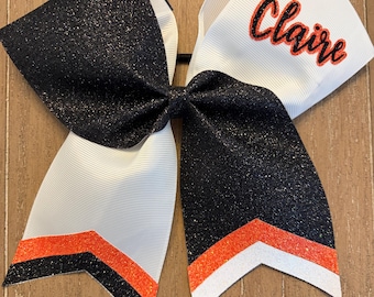 May include: A large hair bow featuring black glitter fabric and white grosgrain ribbon. The bow displays the name Claire in orange script with a black outline. The bottom edges have layered chevron stripes in orange, black, and white glitter.