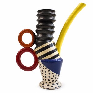 May include: A ceramic pitcher featuring a base with black polka dots on a cream background, a blue triangular section, a middle band with black and white stripes, a tall black ribbed neck, a long yellow spout, and two stacked red and orange rings.