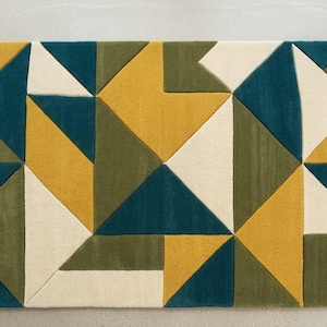 May include: A rectangular area rug featuring a geometric pattern composed of triangles and polygons in shades of mustard yellow, deep teal, olive green, and off-white. The design consists of sharp, clean lines and solid color blocks.