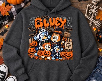Puede incluir: Una sudadera con capucha gris oscuro que presenta un gráfico de cuatro perros de dibujos animados con disfraces de Halloween, incluyendo trajes de esqueleto y bruja, rodeados de calabazas, murciélagos y telarañas, con el texto naranja "BLUEY" y "SPOOKY SEASON".