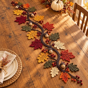 Puede incluir: Un camino de mesa decorativo que presenta una larga enredadera de ganchillo de color marrón oscuro adornada con hojas de colores otoñales en tonos rojo, naranja, amarillo y verde, intercaladas con pequeñas bayas rojas y bellotas de ganchillo.