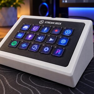 May include: A white Stream Deck control console featuring a black faceplate with fifteen square, backlit LCD keys. Each key displays a unique colorful icon for various software functions, arranged in a three-by-five grid on an angled base.