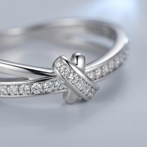Puede incluir: Un anillo de tono plateado con una banda pulida y un diseño cruzado en el centro. La banda y la sección superpuesta están engastadas con una serie de pequeñas piedras redondas y transparentes que ofrecen un aspecto brillante.