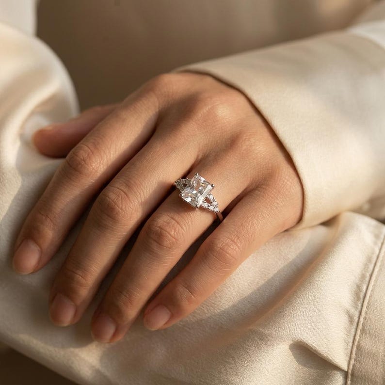May include: A silver-toned ring featuring a large, rectangular, radiant-cut center stone. The band is accented with smaller, round-cut stones arranged in a delicate, open-work pattern on both sides of the central setting, creating a bright, reflective appearance.