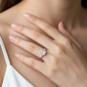 May include: A silver-toned ring featuring a large rectangular center stone flanked by smaller, clear accent stones on a slender band. The ring is worn on a finger, showcasing the faceted design of the stones and the polished metal setting.