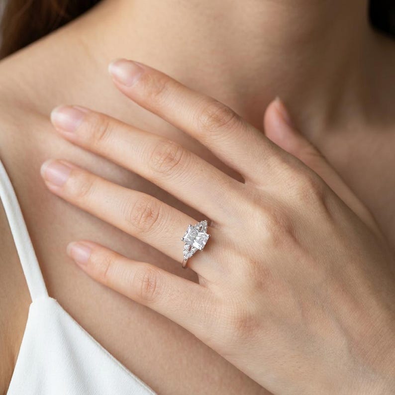 May include: A silver-toned ring featuring a large rectangular center stone flanked by smaller, clear accent stones on a slender band. The ring is worn on a finger, showcasing the faceted design of the stones and the polished metal setting.