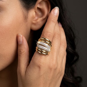 May include: A wide gold ring featuring multiple smooth, polished bands layered together. The center section of the ring is encrusted with numerous small, clear, sparkling stones that create a bright, reflective surface against the gold metal.