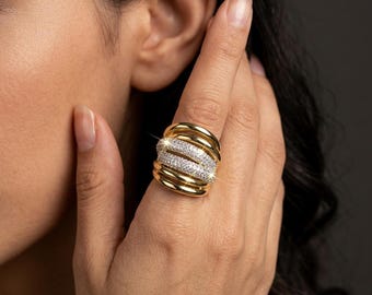 May include: A wide gold ring featuring multiple smooth, polished bands layered together. The center section of the ring is encrusted with numerous small, clear, sparkling stones that create a bright, reflective surface against the gold metal.