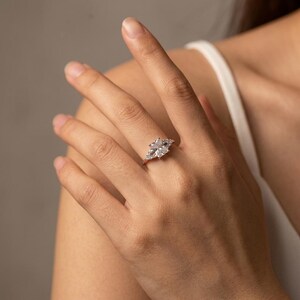 May include: A silver-toned ring featuring a large rectangular radiant-cut center stone flanked by two smaller triangular-cut side stones on each side, set on a thin, polished metal band that encircles the finger with a clear, brilliant sparkle.