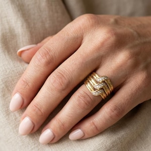 May include: A wide gold band ring featuring a woven or interlocking design. The ring consists of multiple polished gold strands, with one central strand embedded with a row of small, clear, round-cut stones that sparkle against the metal.